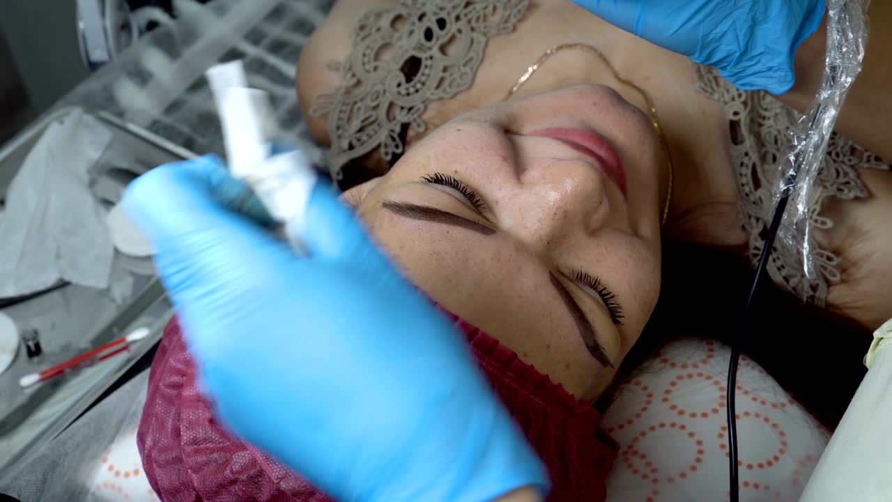 Woman-cosmetician does permanent makeup eyebrow girl in the beauty salon. She applies black paint with a needle on the girl's eyebrows, then wipes it with a swab. Microblading. The view from the top.