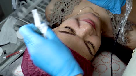 mujer-cosmético hace maquillaje permanente ceja niña en el salón de belleza. ella aplica pintura negra con una aguja en las cejas de la niña, luego lo limpia con un hisopo. microblading. la vista desde la parte superior.