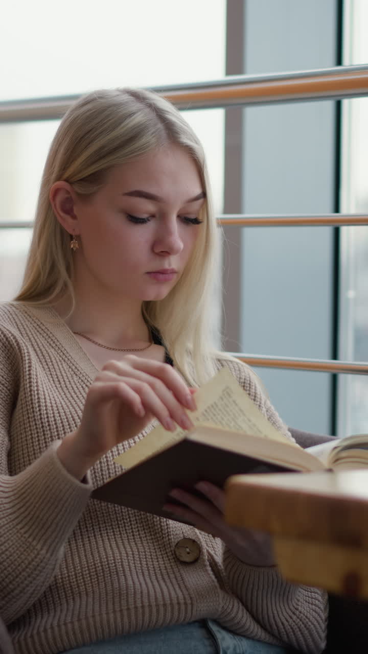 Sinnende Frau blättert in einem Café eine neue Seite um, während sie gedankenversunken ein Buch liest, mit verschwommener Stadtaussicht durch eine Glasscheibe, Kaffeetasse und Telefon auf dem Tisch.