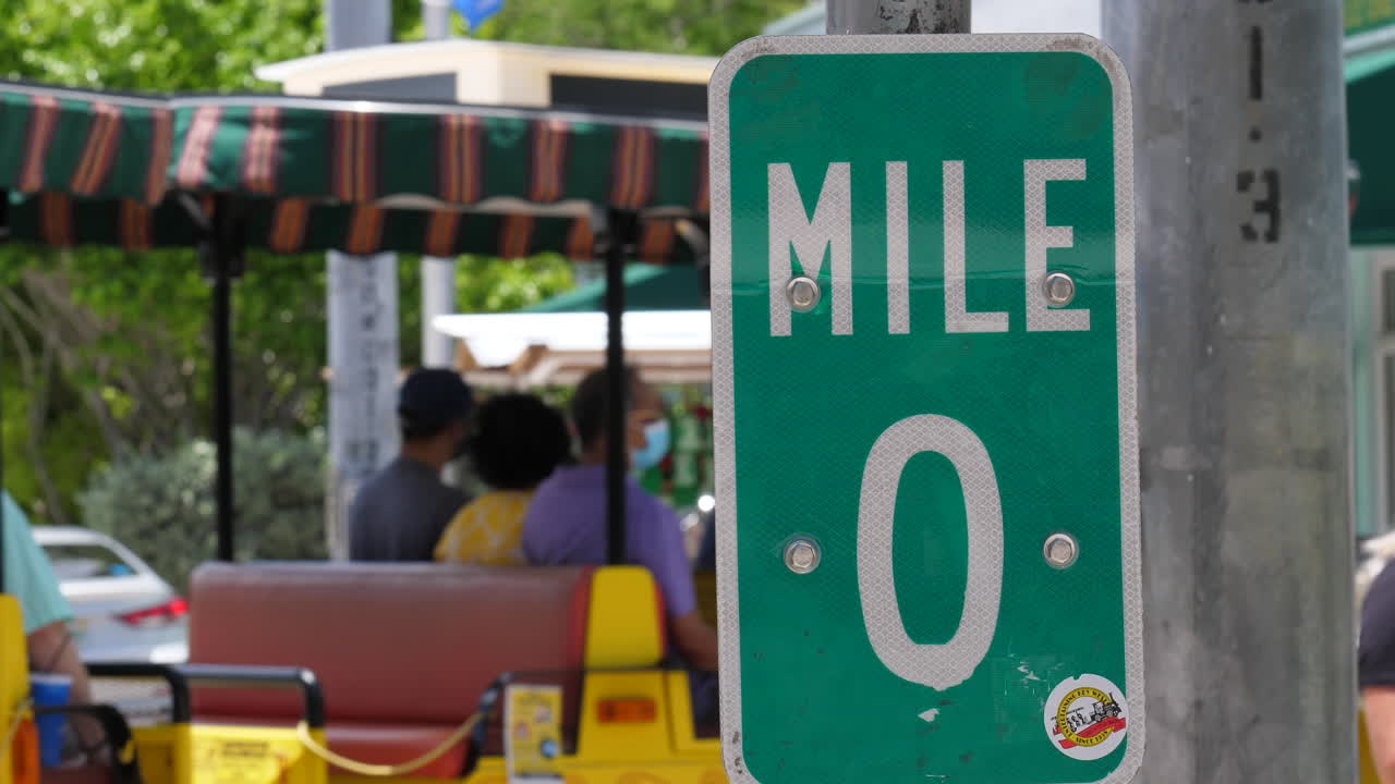 The mile marker 0 sign at the end of US Highway 1 in Key West Florida and a tour train drives by in the background, slow motion.
