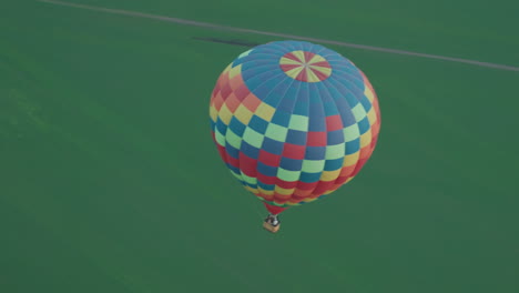 colorful checkered hot air balloon carrying passengers drifts above endless farmland under pastel sky at sunset, aerial perspective showing vivid pattern and sprawling green fields