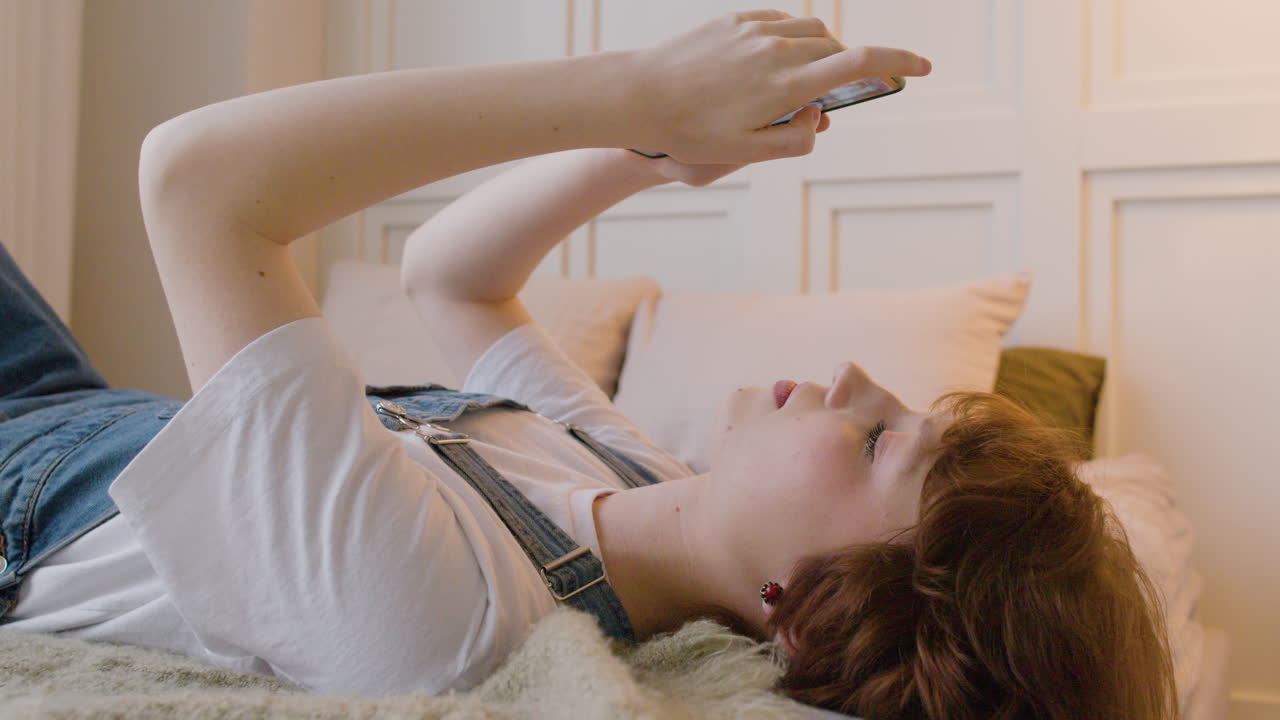 침대에 누워 스마트폰을 사용하여 화면을 탭하고 스크롤하는 소녀의 측면