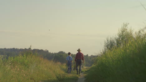 rear view of caucasian grandfather and his little grandson walking together while holding fishing rods in the morning