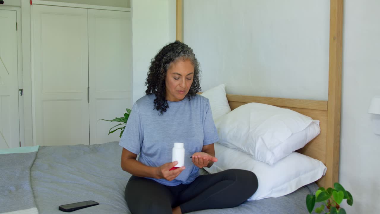 Donna afroamericana seduta a gambe incrociate sul letto che tiene una boccetta di pillole con tappo rosso, con uno smartphone a sinistra.