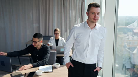young handsome businessman wearing white shirt and trousers stands at window smiling. male and female colleagues working at laptops at their desks.