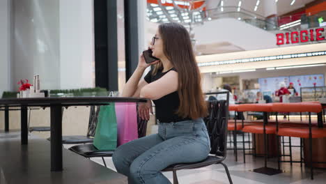 dama sentada en una mesa en un centro comercial ocupado haciendo una llamada telefónica felizmente, bolsas de compras a su lado, personas moviéndose en el fondo, atmósfera comercial moderna con iluminación vibrante