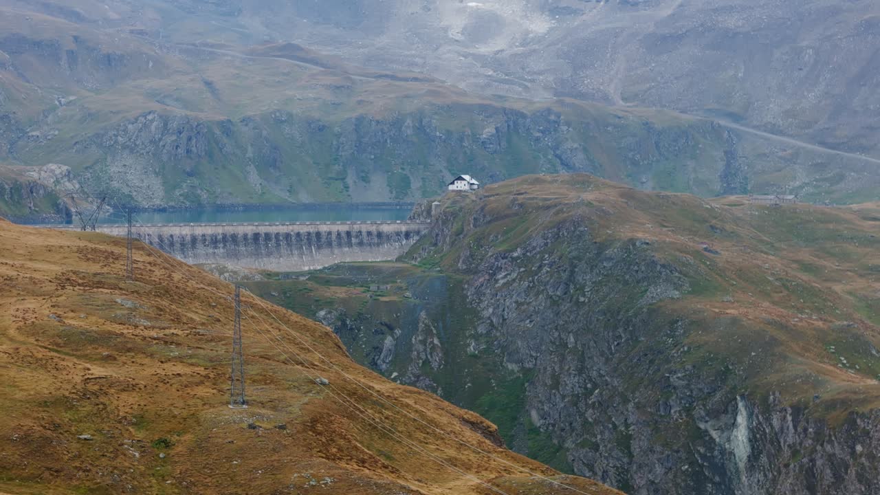 Aerial drone rising shot moving over a house and a vibrant blue reservoir in Cervinia, Italy, framed by dramatic alpine mountains and colorful water tones