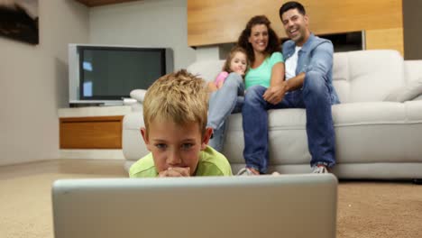 Little-boy-lying-on-floor-using-laptop-with-family-behind-him-on-sofa