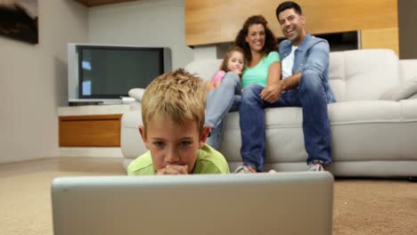 Kleiner-Junge-Liegt-Mit-Laptop-Auf-Dem-Boden,-Familie-Hinter-Ihm-Auf-Dem-Sofa