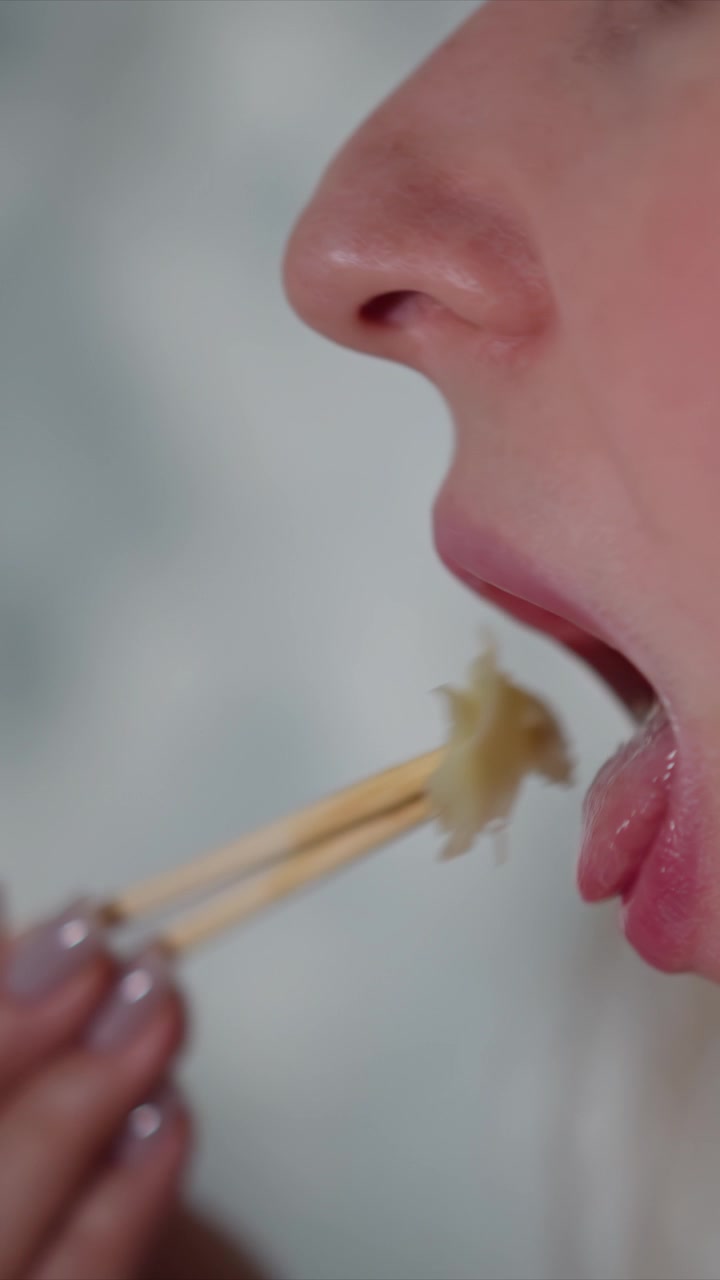 Close up of woman using chopsticks to eat. subject holds wooden utensils near her mouth focusing on action and texture detail against soft background. Lighting is even with no harsh shadows