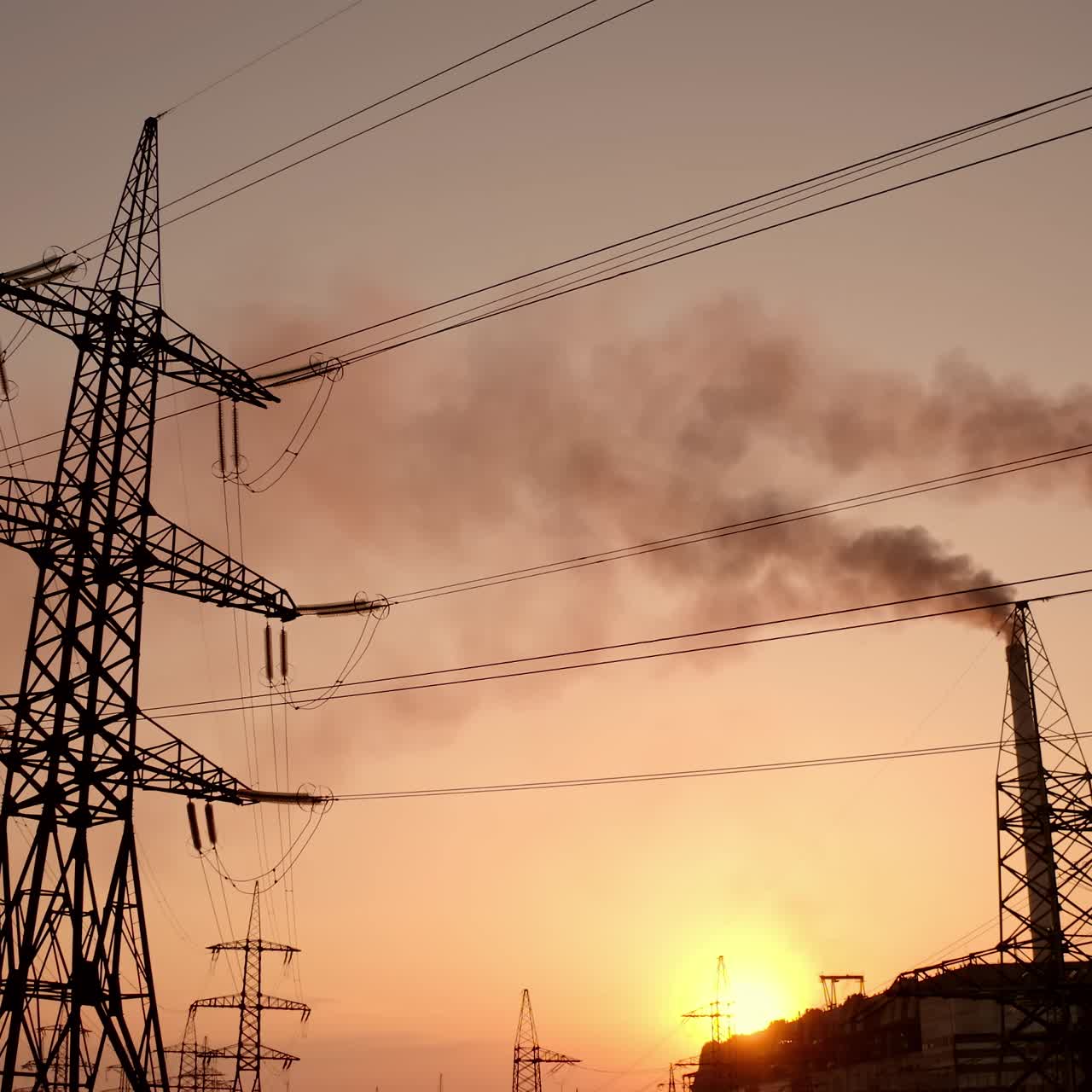 Electricity pylons against smoke stacks at sunset. High-voltage electric towers on the evening sky. Factory pipes with smoke pollute the air. Motion camera down