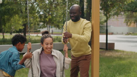 woman swinging on swing in park with help of husband and son