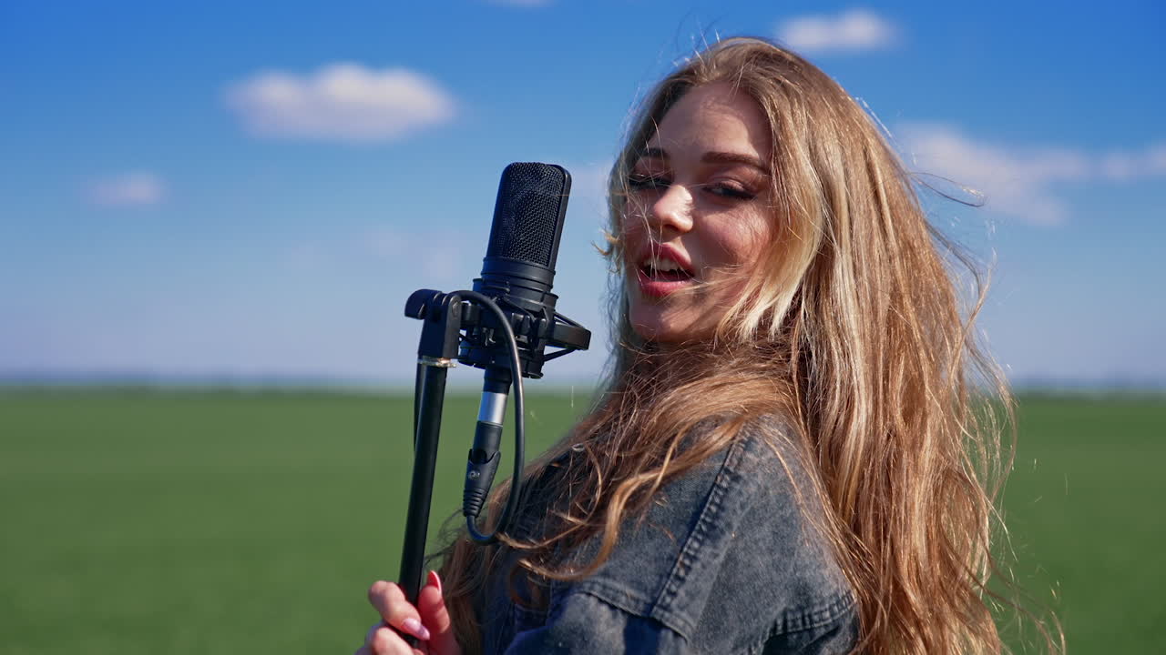Beautiful girl singer in a green field. Attractive blond woman singing into a microphone and energizing looking at the camera.