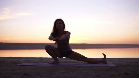 A-sports-brunette-doing-stretching-exercises-on-a-mat-on-the-beach-or-a-lake.-Accelerated-footage.-Front-view