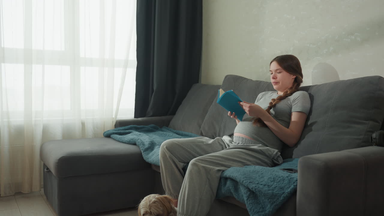 Una donna in attesa si rilassa con il suo cane. Una donna incinta si rilassa comodamente con il suo animale domestico durante una giornata di sole. Una futura mamma gioiosa legge serenamente un libro per bambini mentre il cane riposa nelle vicinanze alla luce del sole.