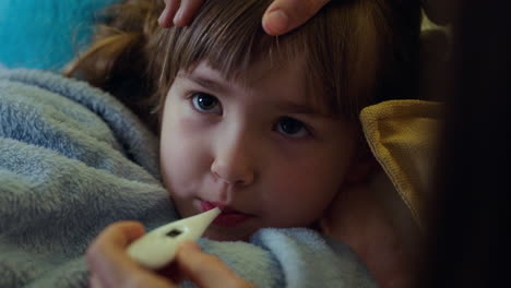 mother caring for sick child lying in bed with flu measuring temperature using thermometer little girl at home with fever mom giving health care