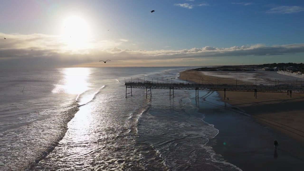 Il tramonto risplende sul mare. Un molo si estende nell'acqua. Le onde scintillano di luce. Le persone camminano felicemente sulla spiaggia. Si godono l'ora d'oro. I gabbiani volano in alto. La scena è pacifica