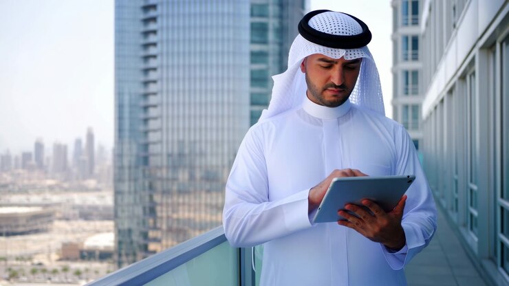 Uomo adulto in thobe bianco e ghutra fantasia con agal nero, in piedi su un moderno balcone di un grattacielo, tiene un tablet con entrambe le mani e tocca lo schermo mentre osserva lo skyline della città sottostante