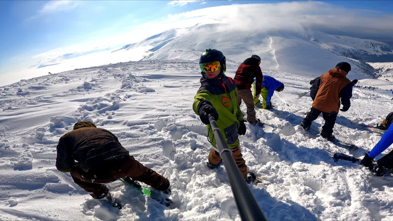 Sportsman in green and black blazer and protective helmet taking selfie with a selfie stick. Man records snowboarders, skiers and grader machine on the snowy mountain.
