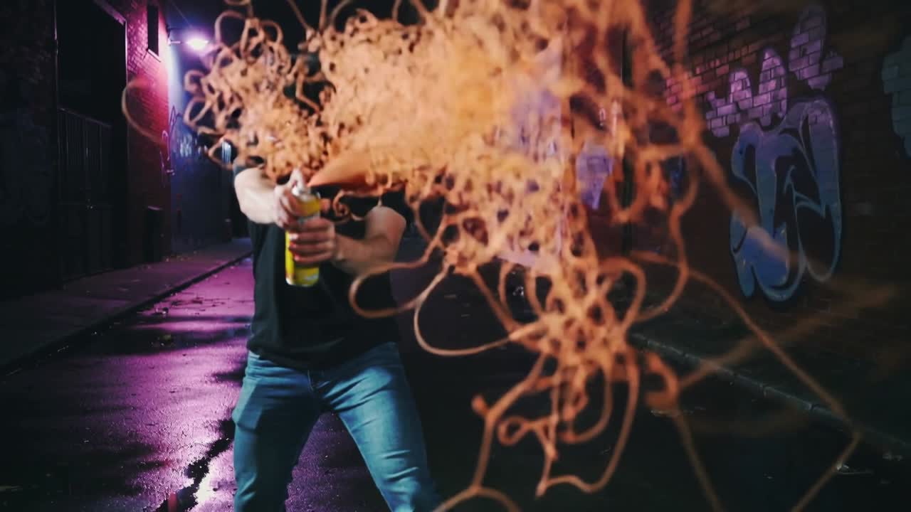 Un uomo che avanza, afferra una bomboletta gialla e preme l'ugello in un vicolo di notte, spruzzando una ragnatela arancione.