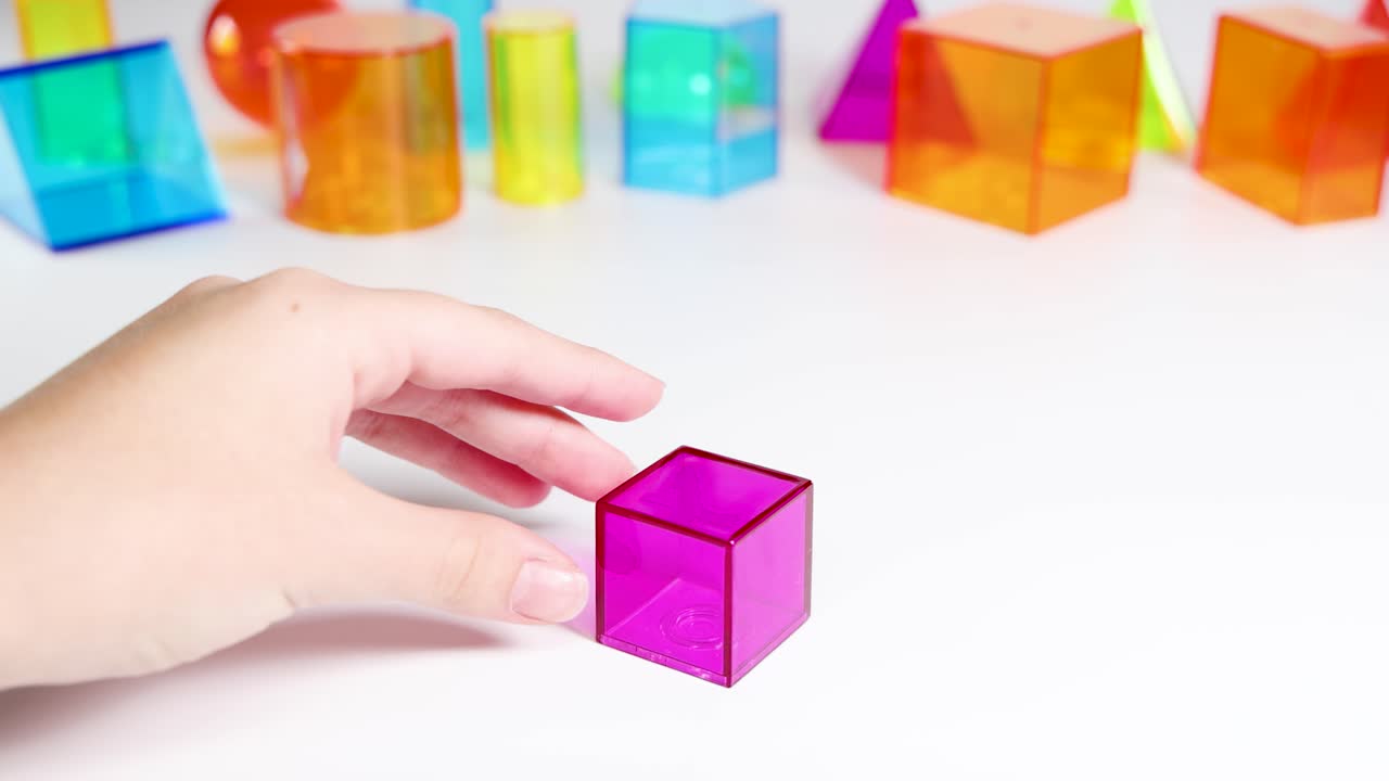 A hand manipulates vibrant 3D geometric shapes on a white surface, highlighting educational and playful interaction