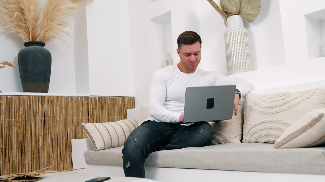 Smiling Caucasian male sits on sofa holding a laptop. Man finishes work, closes his laptop and looks at camera.