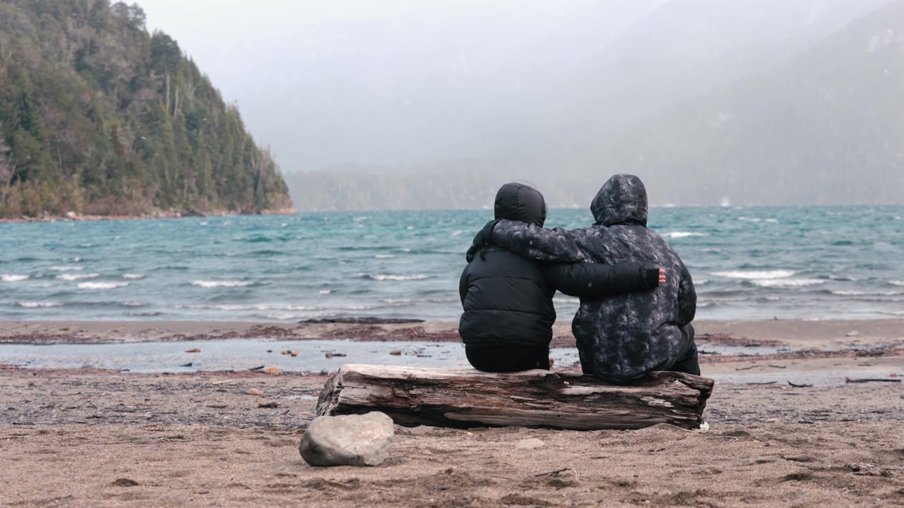 A couple sits on a log by a windy lakeside, embracing each other. The cold weather and choppy waters create a dramatic backdrop. Ideal for themes of support and connection.
