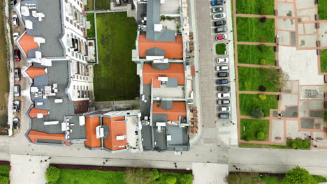 Vista-Aérea-Directamente-Desde-Arriba-De-Una-Hilera-De-Edificios-Con-Techo-De-Terracota-En-Elbląg,-Bordeados-Por-Una-Calle-Con-Autos-Estacionados-Y-Una-Zona-Verde-Con-Césped.