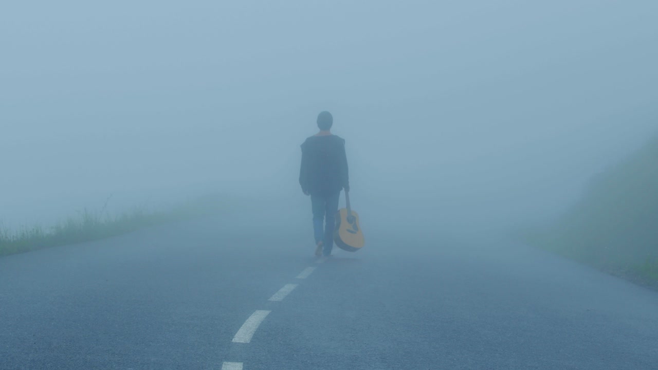 A person is walking on a road in the fog with his guitar