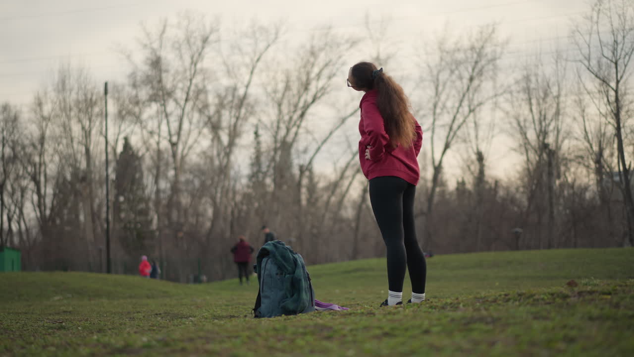 Young Lady Inspects Her Pack Before Exercising, Red Hoodie Woman Examines Her Gear On Grass Before Workout, Young Woman Wearing Red Hoodie Inspects Her Backpack And Exercise Kit Outdoors On Grass