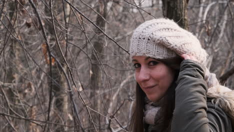 beautiful woman wearing winter clothes in the forest looks and smiles at the camera while touching her hair - medium shot, slow motion