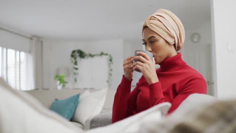 Video-Einer-Gemischtrassigen-Frau-Im-Hijab,-Die-Auf-Dem-Sofa-Sitzt-Und-Kaffee-Trinkt