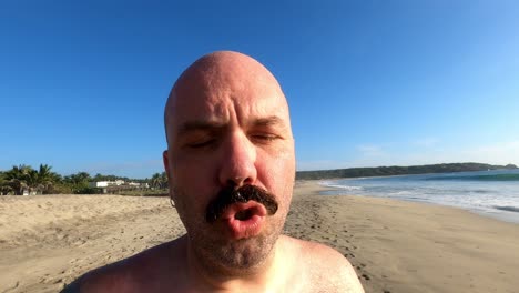 Hombre-Calvo-Caucásico-De-Mediana-Edad-En-La-Playa-Hablando-Frente-A-La-Cámara,-Cerca-De-Un-Turista-De-Vacaciones