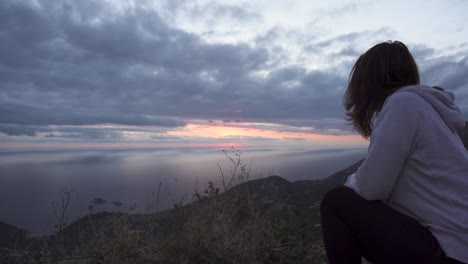 chica en cuclillas en un acantilado alto y ver la puesta de sol a través del cielo nublado sobre el mar adriático-1