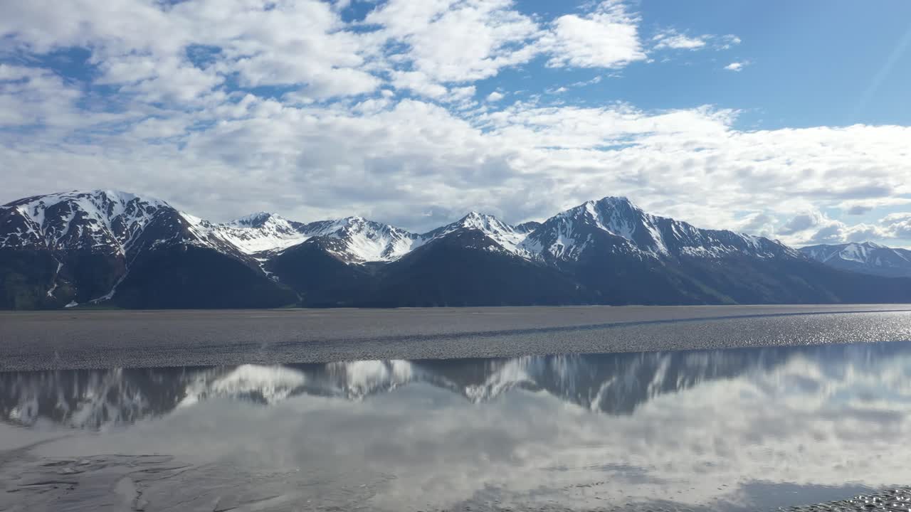드론은 알래스카의 턴가인 팔의 반 ⁇ 이는 물 위로 날아갑니다. 배경에는 케나이 반도의  ⁇ 가치 산맥의 웅장한 눈으로  ⁇ 인 봉우리가 있습니다.