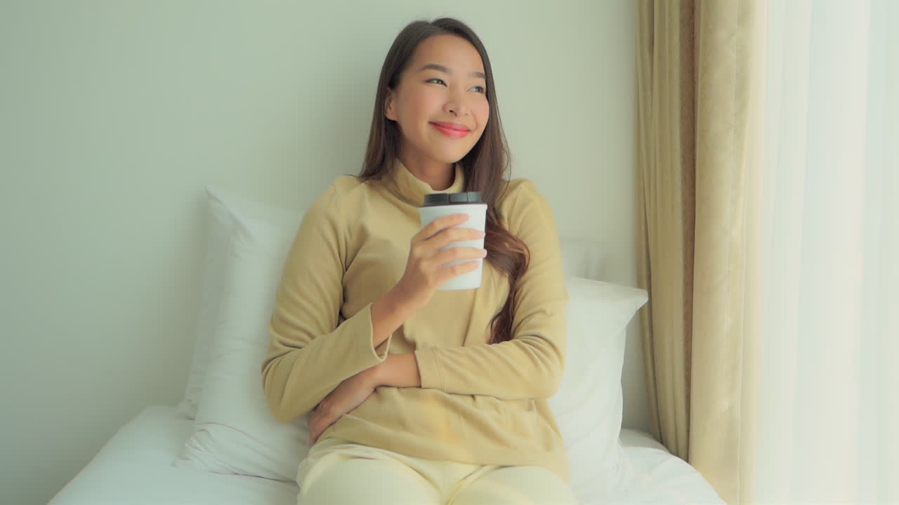 Asian smiling woman drinks coffee sitting on bed while looking out window