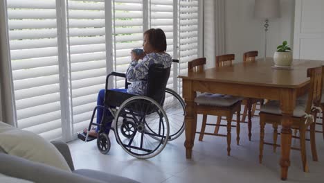 african american senior woman in wheelchair looking out of window at home drinking coffee