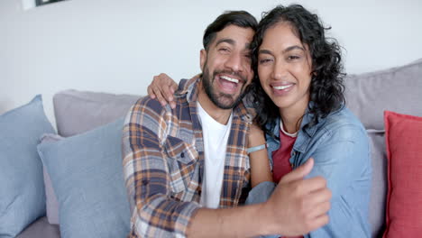 Retrato-De-Una-Feliz-Pareja-Birracial-Sentada-En-Un-Sofá-Abrazándose-Y-Sonriendo-En-Casa,-En-Cámara-Lenta