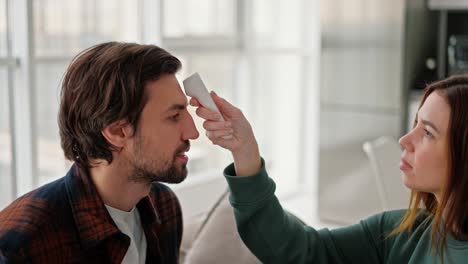 a happy brunette girl in a green sweater measures the temperature of her brunette boyfriend with stubble in an orange-blue shirt on the sofa in a modern apartment