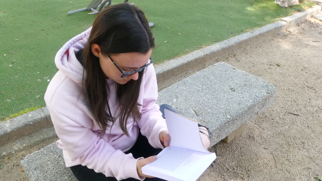 chica joven y linda con gafas leyendo una novela en un banco de piedra en un parque