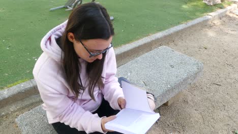 young and cute girl with glasses reading a novel on a stone bench in a park