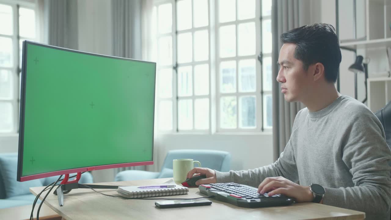 Asian Man In Long Sleeved T-Shirt Typing On Green Screen Desktop Computer While Working At Home.
