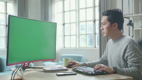 asian man in long sleeved t-shirt typing on green screen desktop computer while working at home.