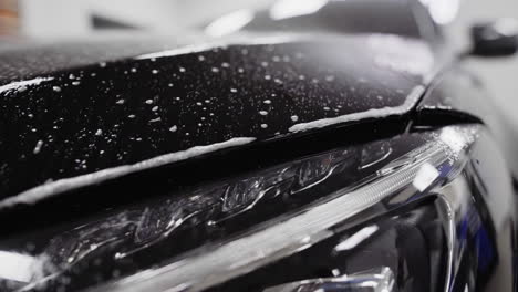 slow motion pan across the front of a black car as sudsy spray wets the hood of the car