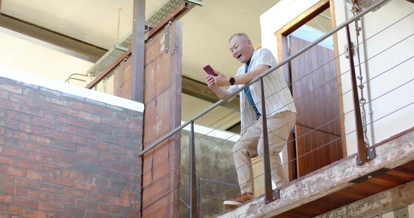Senior Asian man standing near wooden door, leaning on rail and checking red phone, copy space