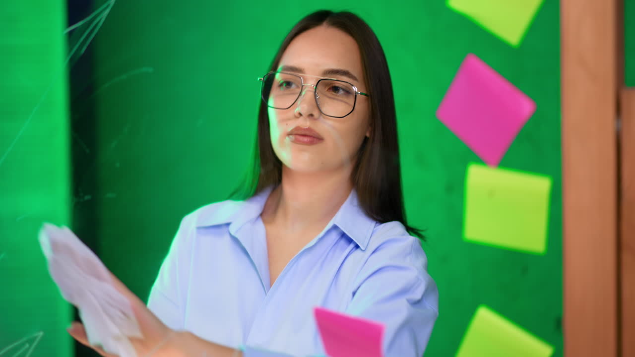 Young woman writes on a green board. A woman in a blue shirt and glasses writes with a marker on a vibrant green board, engaging in a creative activity