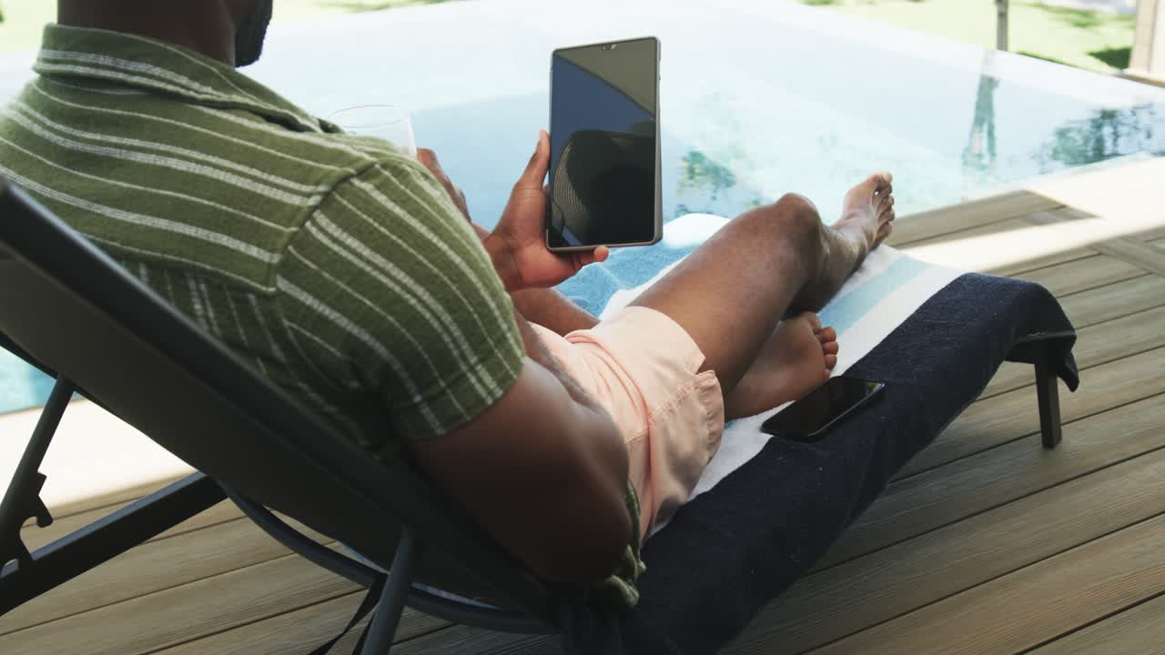 Moden afrikansk mand i grøn skjorte ligger afslappet ved poolen, justerer sin tablet for at læse og nipper til en drink.
