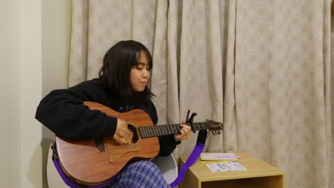 medium shot of pretty, asian girl in a hoodie strumming and playing guitar while singing