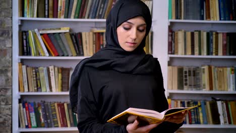 young sweet muslim girl in hijab opens book, reads, religious concept, bookshelf on background