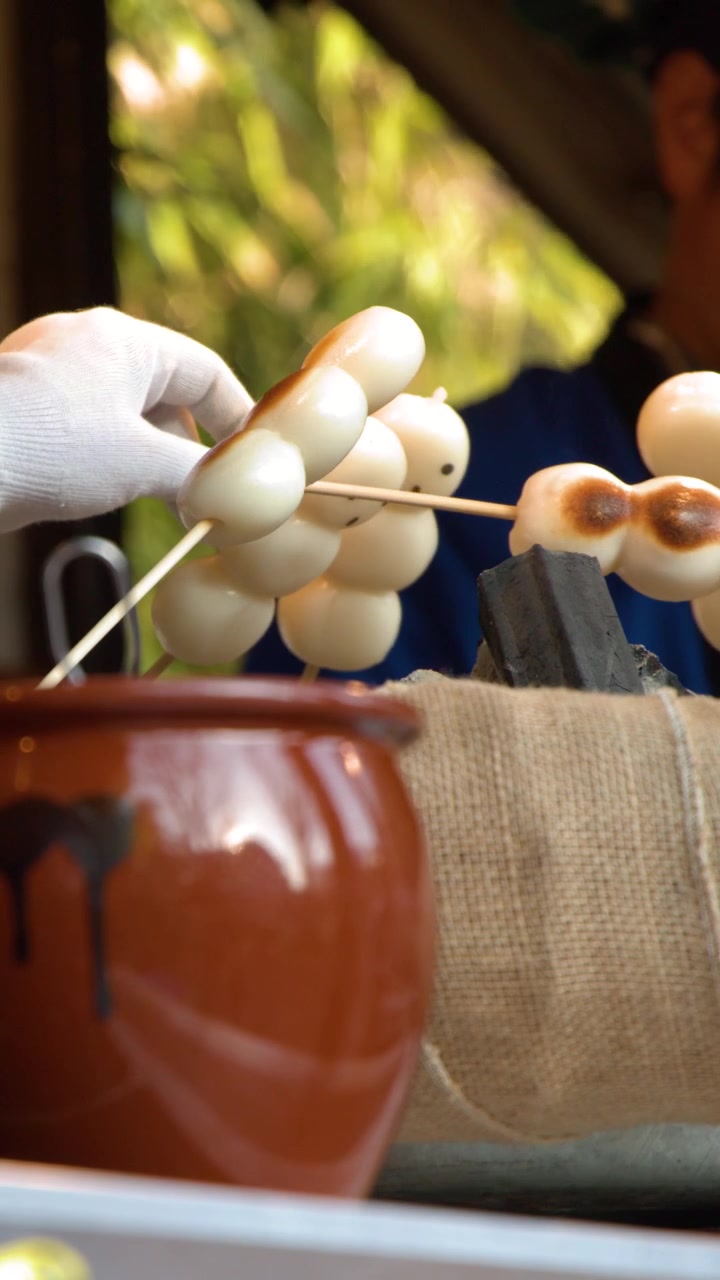Primo piano di un venditore che griglia spiedini di mochi sulla brace, con una pentola di ceramica in primo piano.